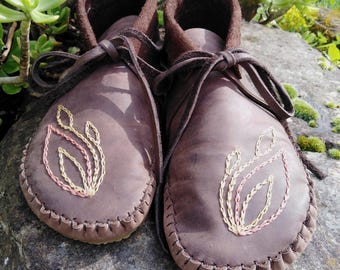 May include: A pair of brown leather moccasins with hand-stitched soles and decorative embroidered leaf designs in yellow, pink, and cream. The moccasins have brown laces and a soft, dark brown interior. The shoes are displayed on a stone surface.