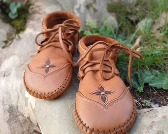 May include: A pair of brown leather baby shoes with a floral design on the toe. The shoes have laces and are stitched around the edges. The shoes are sitting on a tree branch.