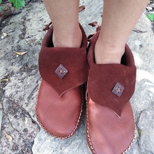 May include: A pair of brown leather moccasins with a folded-over flap and decorative stitching. The flap is a darker shade of brown and features a small, square embellishment. The moccasins are designed with a soft sole.