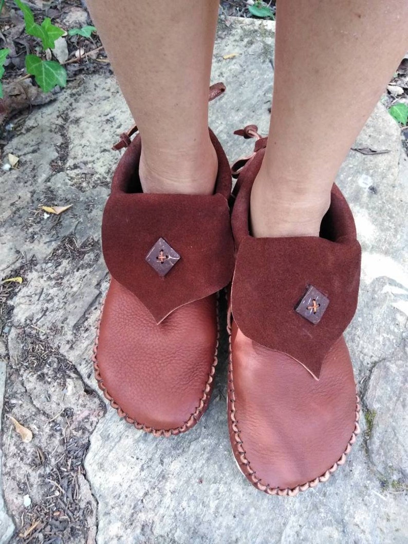 May include: A pair of brown leather moccasins with a folded-over flap and decorative stitching. The flap is a darker shade of brown and features a small, square embellishment. The moccasins are designed with a soft sole.