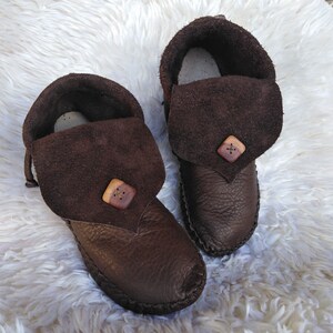 May include: A pair of brown leather moccasin-style slippers. The slippers have a folded-over suede flap secured with a wooden button. The soles are stitched, and the interior appears to be lined with a lighter material. The slippers are on a white, fluffy surface.