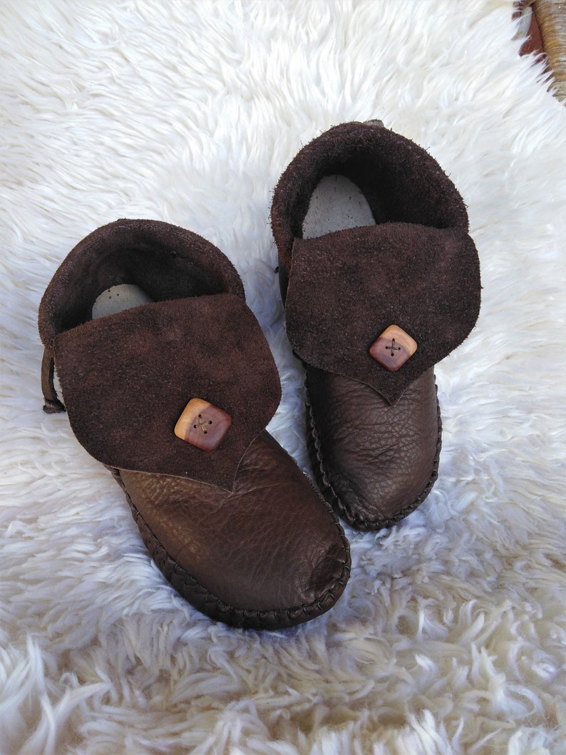 May include: A pair of brown leather moccasin-style slippers. The slippers have a folded-over suede flap secured with a wooden button. The soles are stitched, and the interior appears to be lined with a lighter material. The slippers are on a white, fluffy surface.