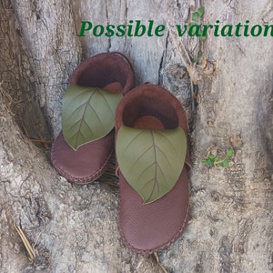 May include: A pair of brown leather baby shoes with a green leaf design on the top. The shoes are made of soft leather and have a stitched sole. The shoes are designed to look like moccasins.