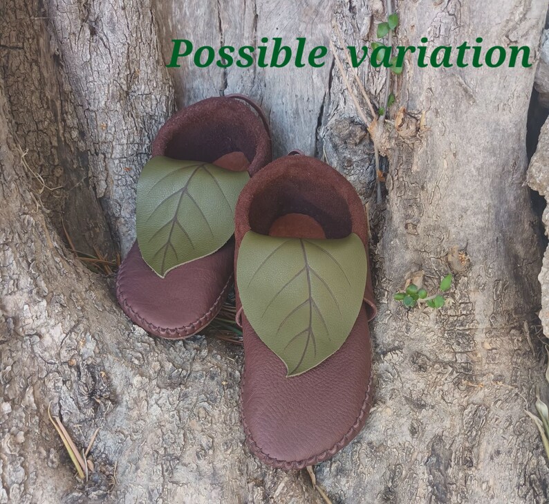 May include: A pair of brown leather baby shoes with a green leaf design on the top. The shoes are made of soft leather and have a stitched sole. The shoes are designed to look like moccasins.