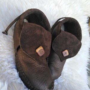 May include: A pair of brown leather moccasin-style shoes. The shoes have a dark brown, textured leather body with a stitched sole. Each shoe features a suede flap secured with a wooden button. A leather strap extends from the back of each shoe.