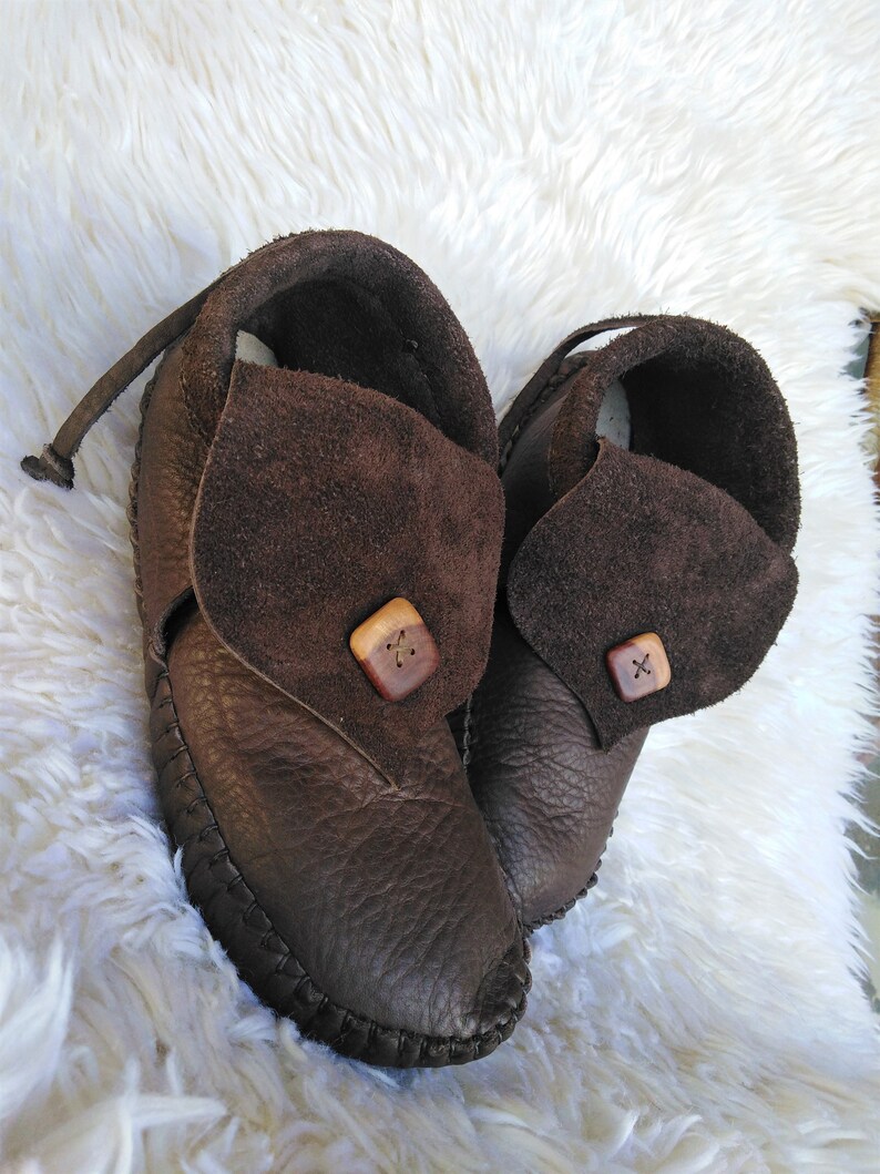 May include: A pair of brown leather moccasin-style shoes. The shoes have a dark brown, textured leather body with a stitched sole. Each shoe features a suede flap secured with a wooden button. A leather strap extends from the back of each shoe.