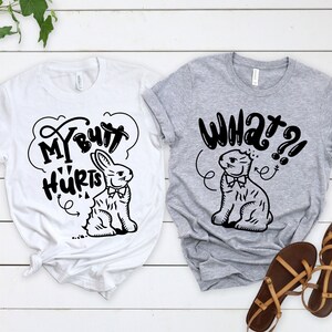 May include: Two t-shirts with a black and white illustration of a bunny rabbit. The white t-shirt says "My Butt Hurts" and the gray t-shirt says "What?!".