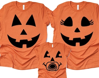 May include: Three orange t-shirts with black jack-o'-lantern faces. Two adult shirts have triangular eyes and a wide smile. The child's shirt has a pacifier design. Perfect for Halloween family costumes.
