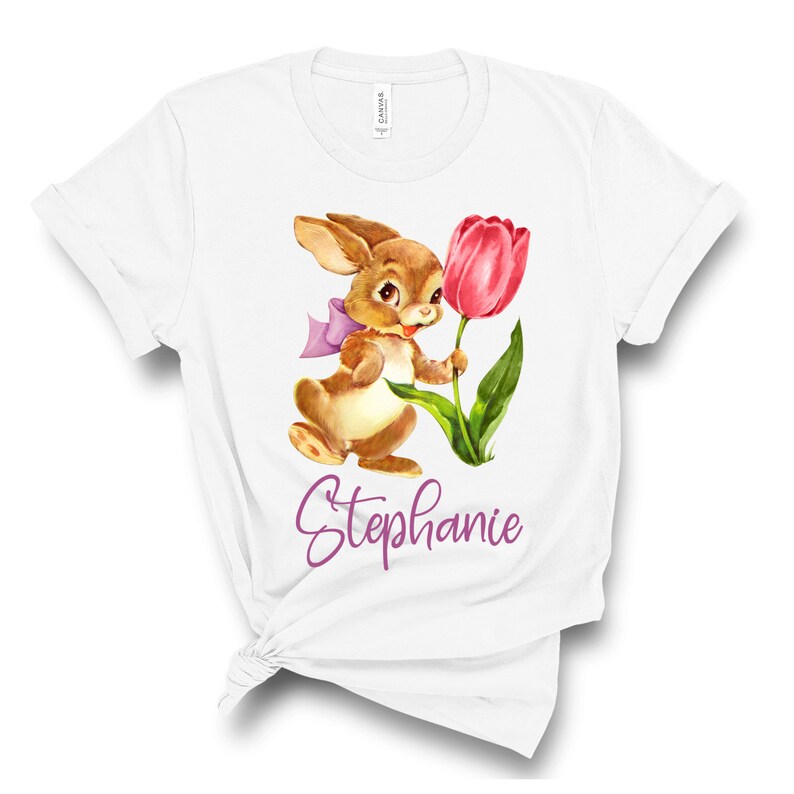 Pu&ograve; includere: Maglietta bianca con un coniglio marrone che tiene un tulipano rosa. Il coniglio ha un nastro viola intorno al collo. Il testo "Stephanie" &egrave; stampato in viola sotto il coniglio.