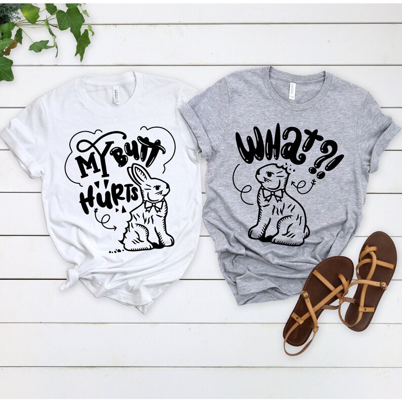 May include: Two t-shirts with a black and white illustration of a bunny rabbit. The white t-shirt says "My Butt Hurts" and the gray t-shirt says "What?!".