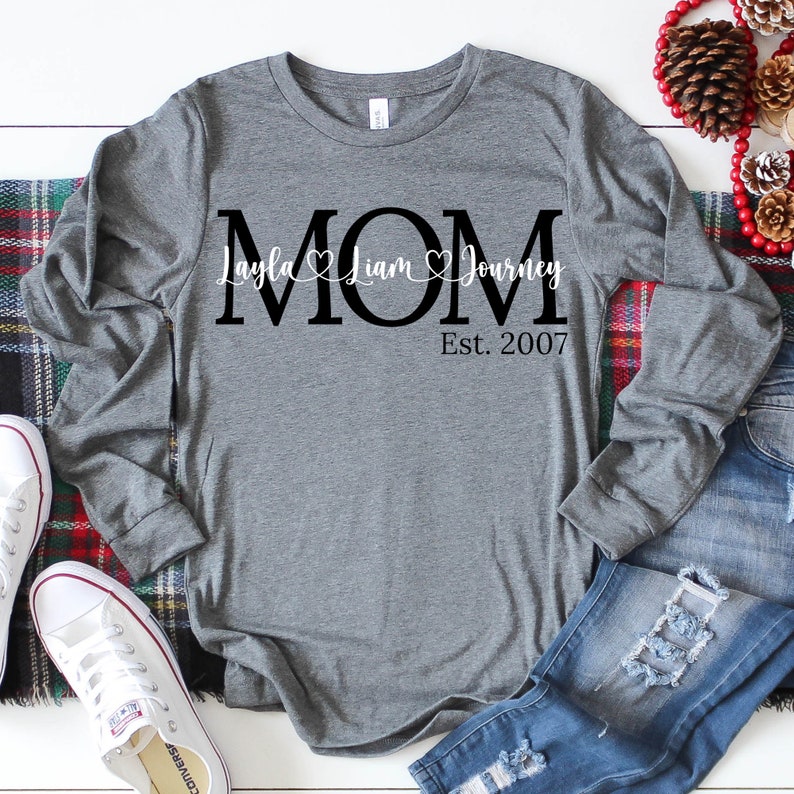 Op de afbeelding: Grijs longsleeve shirt met zwarte tekst die "MOM" zegt met de namen "Layla", "Liam" en "Journey" en de tekst "Est. 2007" eronder.