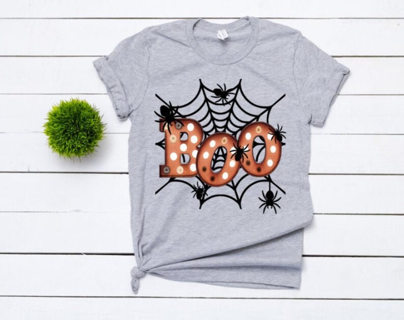 K&ouml;nnte beinhalten: Hellgraues T-Shirt mit einem Halloween-Motiv. Das Design zeigt das Wort "BOO" in orangefarbenen Buchstaben mit wei&szlig;en Punkten, umgeben von einem schwarzen Spinnennetz und schwarzen Spinnen. Links befindet sich eine kleine gr&uuml;ne Dekokugel.
