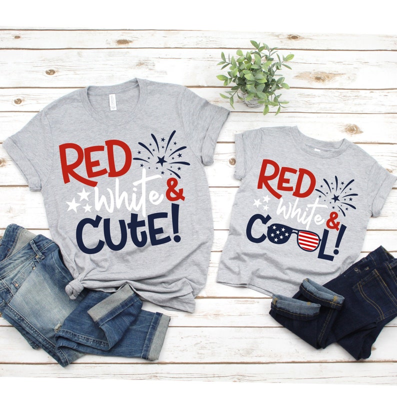 May include: Two gray t-shirts with red, white, and blue text and graphics. The larger shirt says "Red White & Cute!" with a firework graphic. The smaller shirt says "Red White & Cool!" with a graphic of sunglasses with an American flag design.