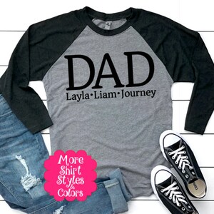 May include: A gray and black baseball tee shirt with the word "DAD" in black lettering and the names "Layla, Liam, Journey" underneath. The shirt is on a white wood background with a pair of blue jeans and black sneakers.