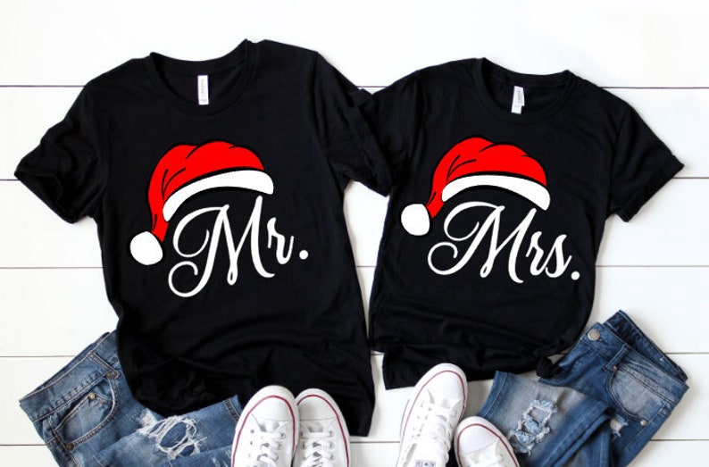 Puede incluir: Dos camisetas negras con un dise&ntilde;o de gorro de Pap&aacute; Noel rojo y blanco. La camiseta de la izquierda dice "Mr." y la camiseta de la derecha dice "Mrs."