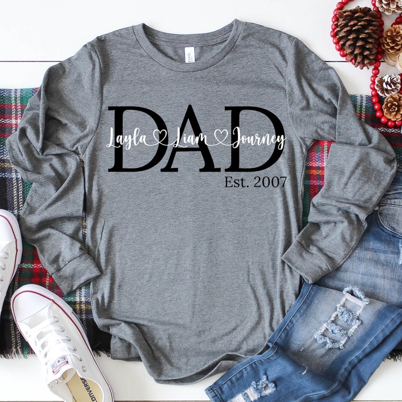 May include: Gray long-sleeve shirt with the word "DAD" in large black letters. The names "Layla", "Liam", and "Journey" are written in smaller letters above the word "DAD" with hearts between each name. The text "Est. 2007" is written below the word "DAD".