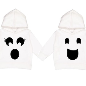 May include: Two white hooded sweatshirts with ghost faces. One has surprised eyes and mouth, the other has a smiling face. The sweatshirts are perfect for Halloween or costume parties.