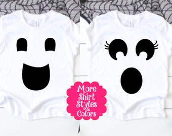 May include: Two white t-shirts with black ghost face designs. One shirt has a smiling ghost face, and the other has a surprised ghost face with eyelashes. A pink flower-shaped graphic with text is in the lower left corner.