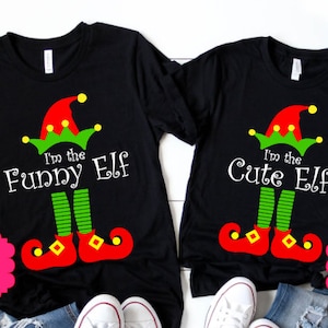May include: Two black t-shirts with elf-themed designs. One shirt says "I'm the Funny Elf" and the other says "I'm the Cute Elf". Each shirt features a red and green elf hat and shoes. The shirts are displayed on a white background.