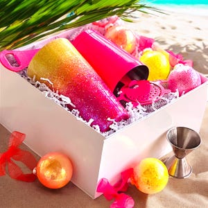 Pode incluir: Uma caixa de presente branca cheia de suprimentos para fazer coquetéis. Inclui um copo degradê rosa e amarelo, uma coqueteleira rosa brilhante, um coador e um jigger. A caixa também contém enfeites decorativos e é forrada com papel branco.