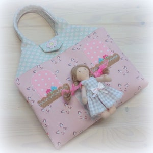 May include: A pink tote bag with a cat face pattern and a light blue floral patterned top. The bag features two window appliqu&eacute;s with pink polka dots and a doll in a checkered dress.