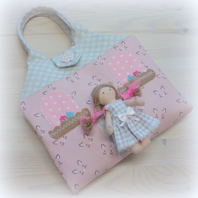 May include: A pink tote bag with a cat face pattern and a light blue floral patterned top. The bag features two window appliqu&eacute;s with pink polka dots and a doll in a checkered dress.