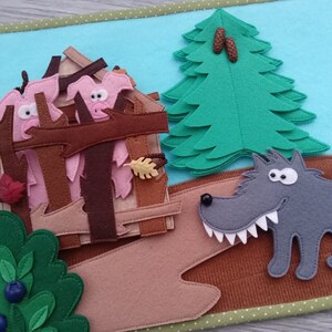 May include: A felt quiet book page featuring a scene with a gray wolf, a house with pink pig characters, a green tree, and a bush with blue berries. The background is light blue, and the edges are green.