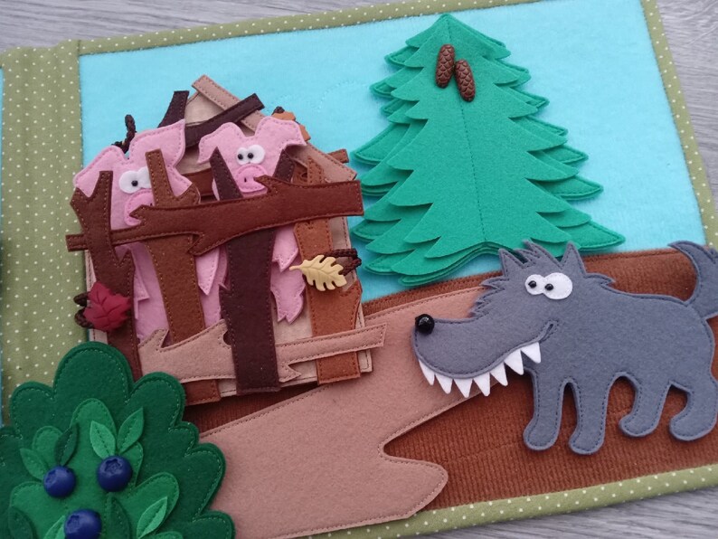 May include: A felt quiet book page featuring a scene with a gray wolf, a house with pink pig characters, a green tree, and a bush with blue berries. The background is light blue, and the edges are green.