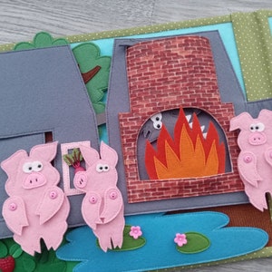 May include: A felt quiet book page featuring a scene from the Three Little Pigs. The page shows three pink pigs, a brick chimney with a fire, and a gray house. The scene includes a pond with lily pads and flowers.