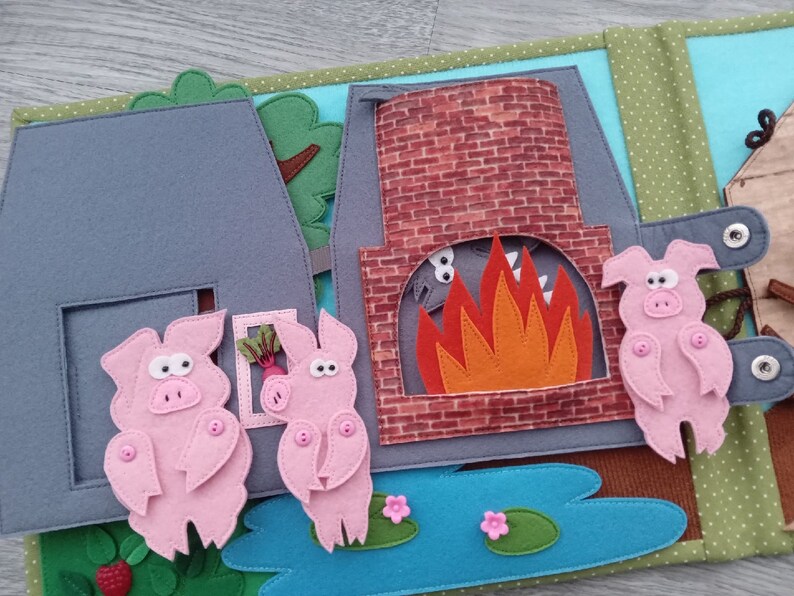May include: A felt quiet book page featuring a scene from the Three Little Pigs. The page shows three pink pigs, a brick chimney with a fire, and a gray house. The scene includes a pond with lily pads and flowers.