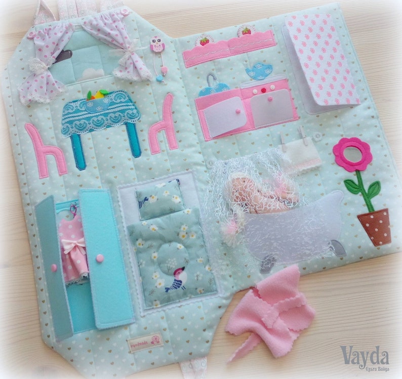 May include: A handmade, soft fabric activity book with a house theme. The open book features a kitchen, bedroom, and bathroom scenes with felt details. Includes a pink dress in a closet, a doll in a bathtub, and a flower pot.