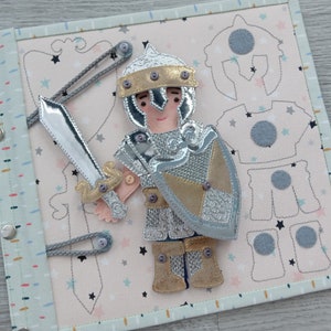 May include: A fabric activity book featuring a knight with a silver sword and shield. The knight is wearing silver and gold armor. The background is light pink with star and shield patterns. The book has a light blue border and silver snaps.