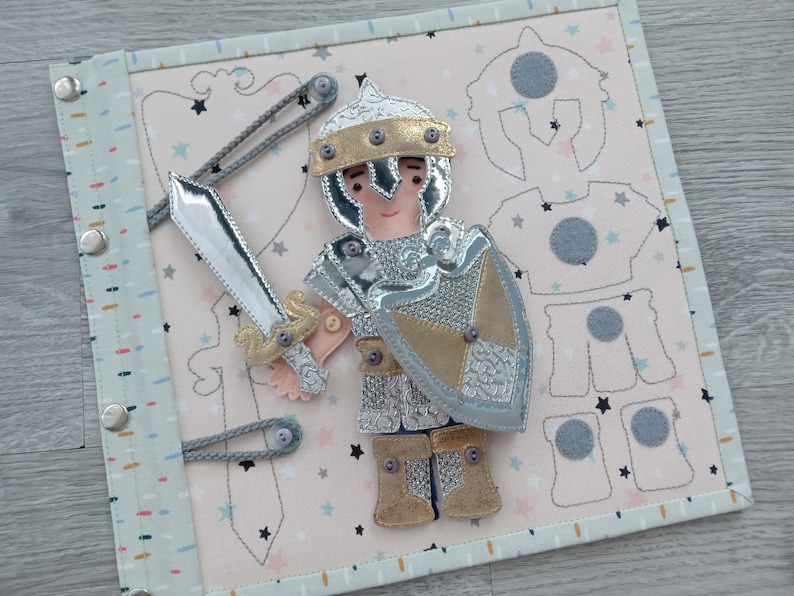 May include: A fabric activity book featuring a knight with a silver sword and shield. The knight is wearing silver and gold armor. The background is light pink with star and shield patterns. The book has a light blue border and silver snaps.