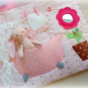 May include: A pink and white quilted fabric activity mat with a bunny in a pink polka-dot tub. Features include a flower with a mirror, a felt bee, a clothesline with a towel, and a lace curtain. Gold heart accents.