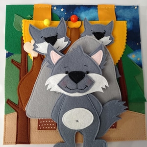 May include: A handmade felt quiet book with a wolf theme. The book features a yellow flap with three wolf faces, a forest scene, and a night sky background. The main wolf is grey with white and pink accents.