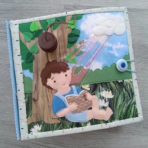 Könnte beinhalten: Handgemachtes Filz-Quiet-Book mit einer Naturszene. Die Seite zeigt einen Jungen, der unter einem Baum liest, mit einer Wolke und einem Regenbogen. Das Buch hat einen blauen Knopfverschluss und einen gemusterten Rand. Die Szene umfasst Filzblätter und -blumen.