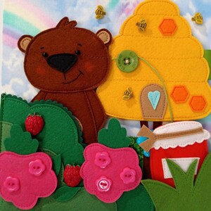 May include: A colourful felt craft scene depicting a brown bear, a yellow beehive with bees, and pink flowers with button centres. A red jar of honey and strawberries are also present. The background shows a rainbow and blue sky.