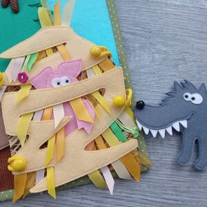 May include: A felt craft depicting a scene from the Three Little Pigs. A pink pig peeks out from a straw house, with a gray wolf nearby. The house is tan with colorful ribbons and yellow accents. Green and blue felt trees are in the background.