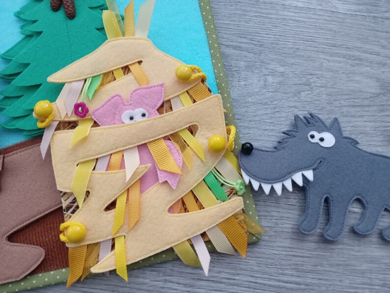 May include: A felt craft depicting a scene from the Three Little Pigs. A pink pig peeks out from a straw house, with a gray wolf nearby. The house is tan with colorful ribbons and yellow accents. Green and blue felt trees are in the background.