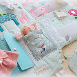 May include: A soft, quilted fabric activity book with various interactive elements. Features a doll in a bed, a pink dress, and a drawstring bag. The book has a light blue background with pink and turquoise accents.