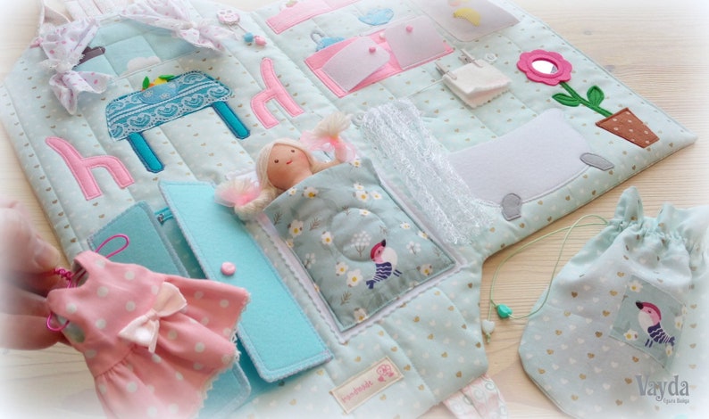 May include: A soft, quilted fabric activity book with various interactive elements. Features a doll in a bed, a pink dress, and a drawstring bag. The book has a light blue background with pink and turquoise accents.