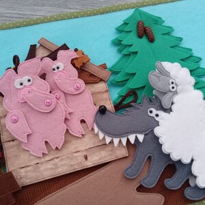 May include: A felt quiet book page featuring a scene from the Three Little Pigs story. Pink pigs stand near a brown house, while a gray wolf menaces a sheep near a green tree. The background is blue.
