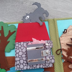 May include: A felt quiet book page depicting a scene from the Three Little Pigs. The page shows a gray wolf, a red-roofed stone house, and a tree. The house has a gray door with silver clasps, and pink pig faces are visible inside.