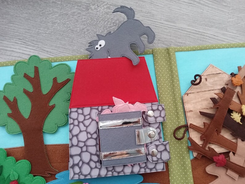 May include: A felt quiet book page depicting a scene from the Three Little Pigs. The page shows a gray wolf, a red-roofed stone house, and a tree. The house has a gray door with silver clasps, and pink pig faces are visible inside.
