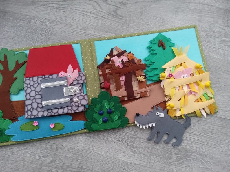 May include: A felt storybook open to a scene from the Three Little Pigs. The pages show the pigs' houses, a pond, trees, and a wolf. The houses are made of brick, wood, and straw. The wolf is gray and has sharp teeth.