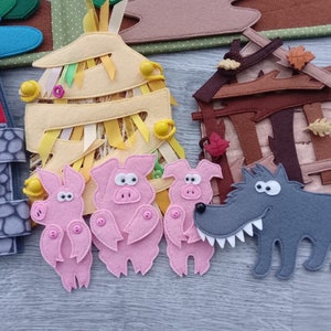 May include: A felt craft depicting a scene from the Three Little Pigs story. The scene includes three pink pigs, a gray wolf, and three houses made of straw, wood, and brick. The houses have colorful details and ribbons.