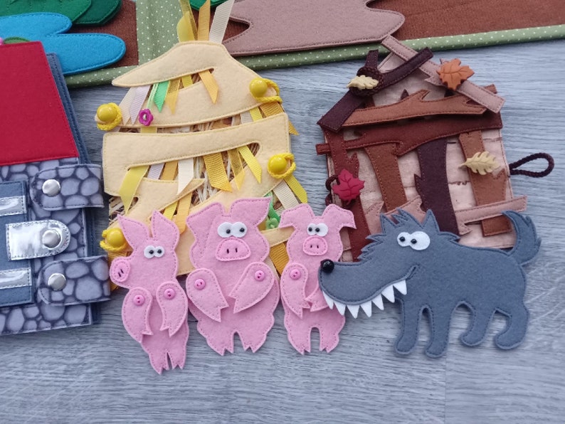 May include: A felt craft depicting a scene from the Three Little Pigs story. The scene includes three pink pigs, a gray wolf, and three houses made of straw, wood, and brick. The houses have colorful details and ribbons.