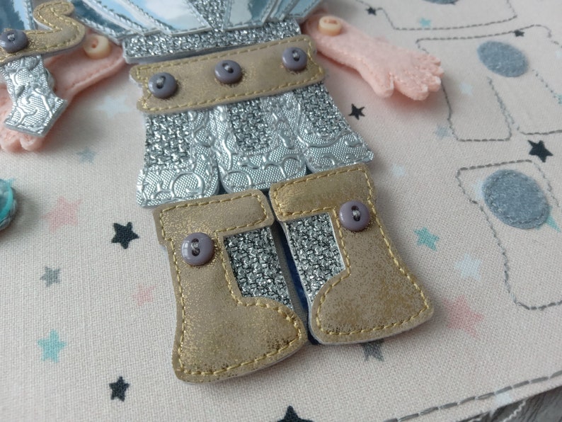 May include: Close-up of a felt doll with silver armor and gold boots. The doll has a light pink arm and is decorated with small gray buttons. The background is a light pink fabric with star and boot patterns.