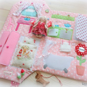 May include: A pink and white polka-dotted fabric activity mat with various interactive elements. Features include a doll, miniature furniture, a wardrobe, a bathtub, and a dog toy. The mat is designed for children and includes a variety of textures and shapes.