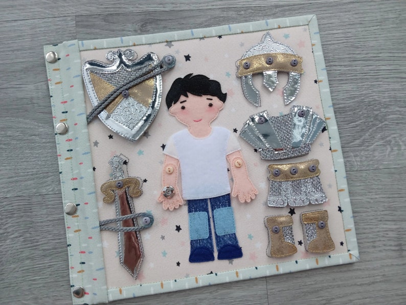May include: A fabric activity board featuring a felt figure with removable knight armor pieces. The armor includes a helmet, chest plate, skirt, boots, shield, and sword. The background is a light pink with star shapes.